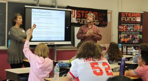 A student in the front row raises her hands as two adults speak at the front of the classroom