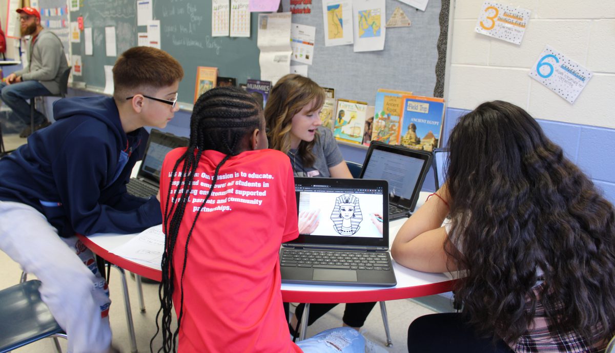 Students sitting at a table look at a teacher's screen