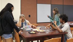 Two adults sit at a table as one girl stands near leaning on the table and another girl walks by 