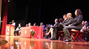 A student speaks on stage as administrators and staff members watch