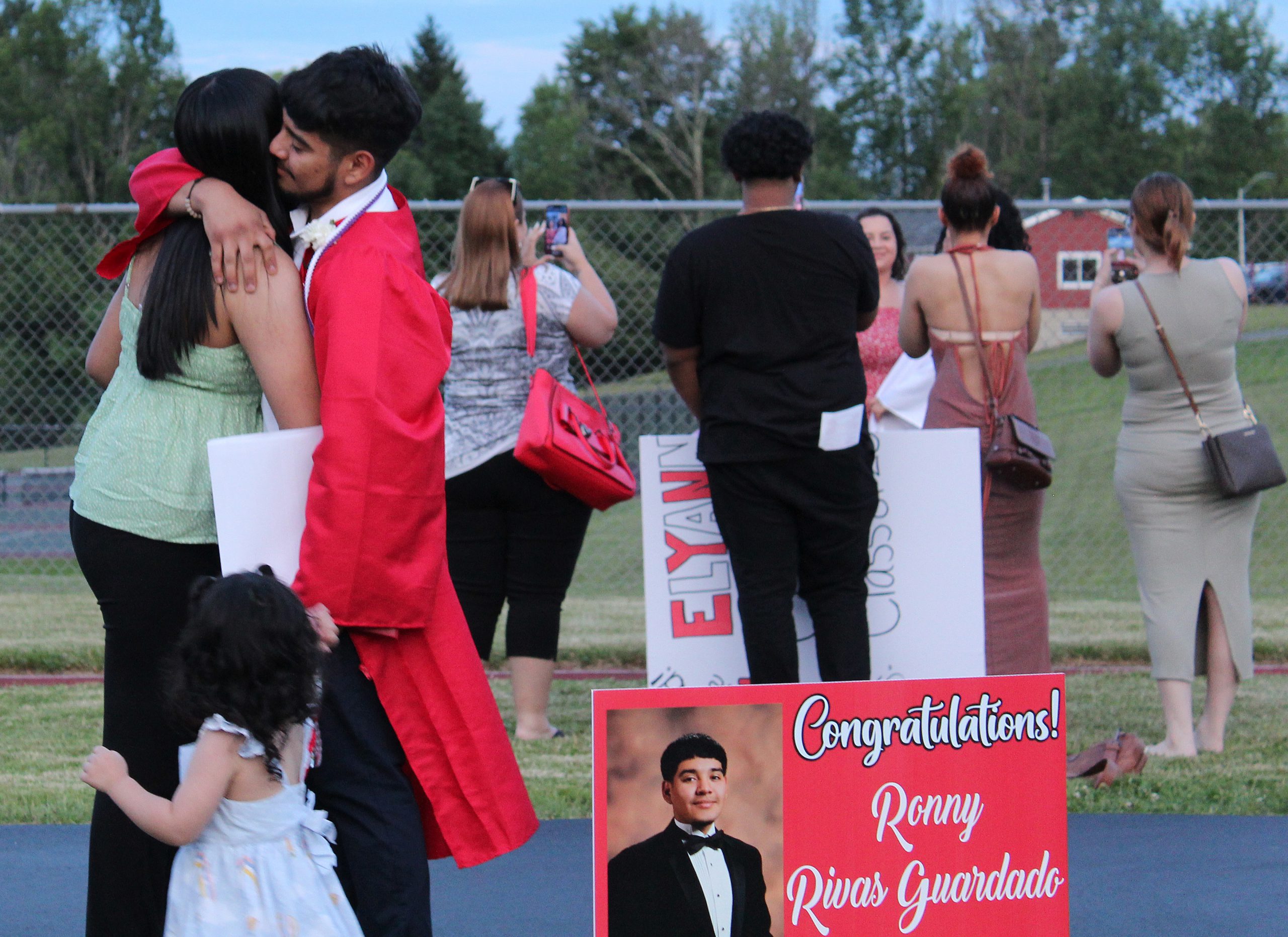 IMG_7748 A graduate hugs an adult with a sign graduation sing in front of them as another group prepares to take photos in the back ground