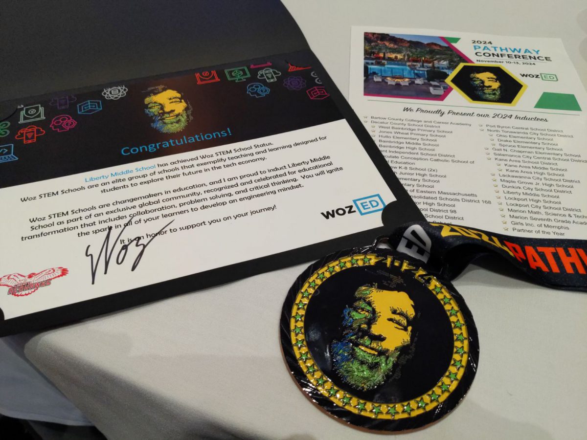 A certificate and medal recognizing Liberty Middle School as Woz Ed Pathway school sits along with an agenda from a conference sits on a table.