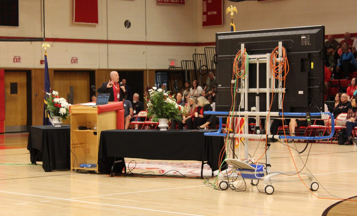 A man talks in front of a crowd with tables and screens behind him in a gymnasium