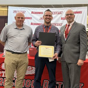 A man holds a certificate and a a piece of paper with a pin on it flanked by two men.