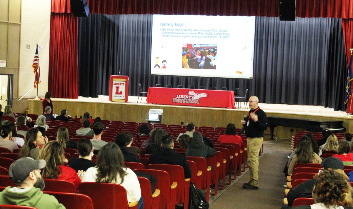A man speaks in an auditorium with a large screen behind him