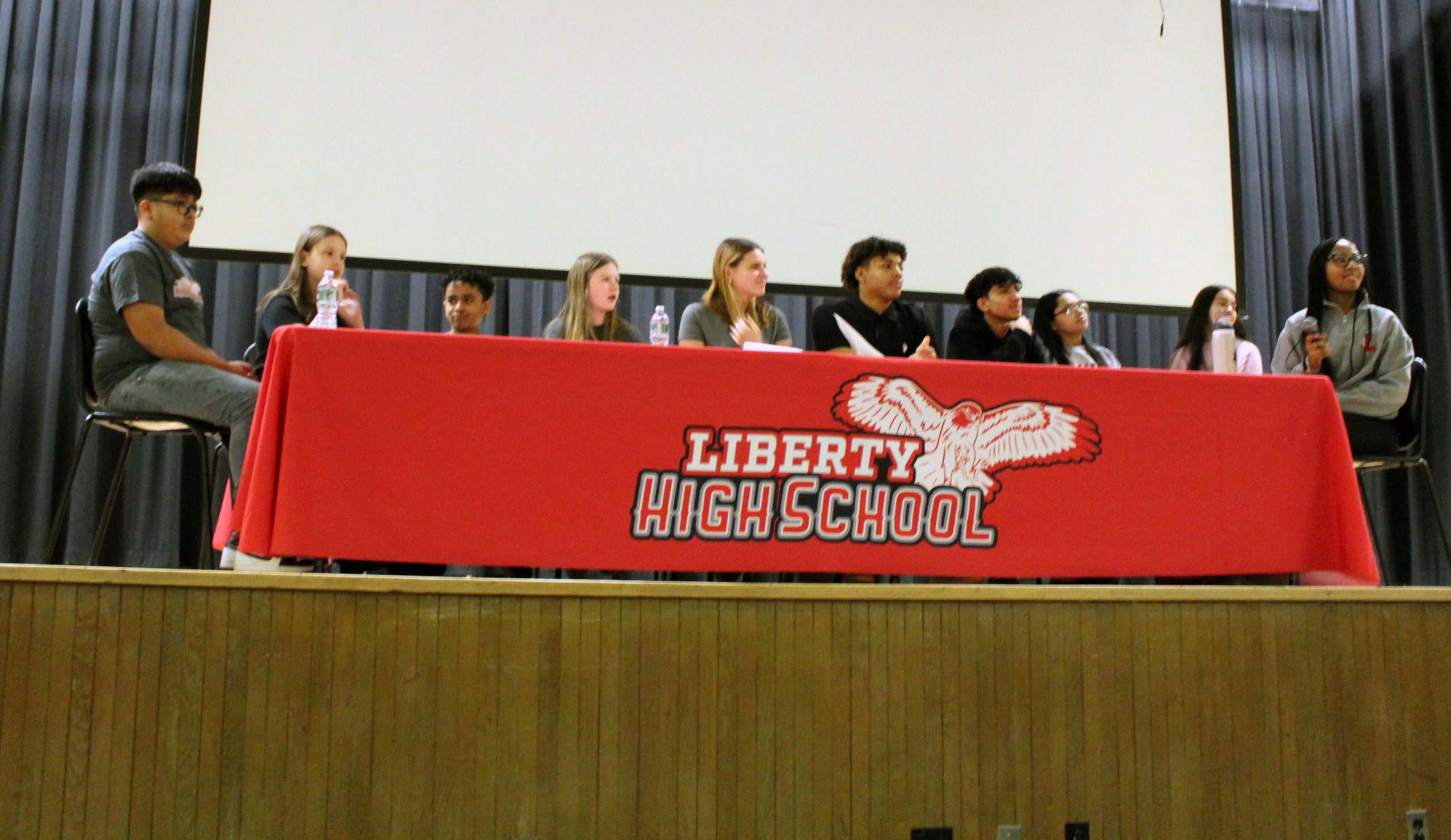 10 students sit on stage at a table with a red tablecloth with the Liberty High School logo on it.