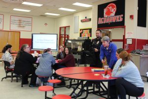 A person seated at a table reads from a note on book into a microphone held by another adult as a police officer holds a book bag.