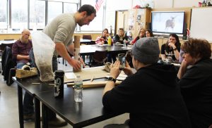 A man shapes a clay cylinder around a tube as others seated at tables watch and one record it on a phone. 