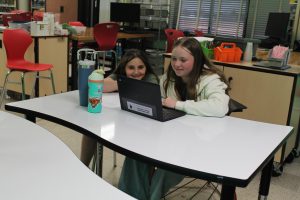 Two students sit at a desk watching a video on a Chromebook