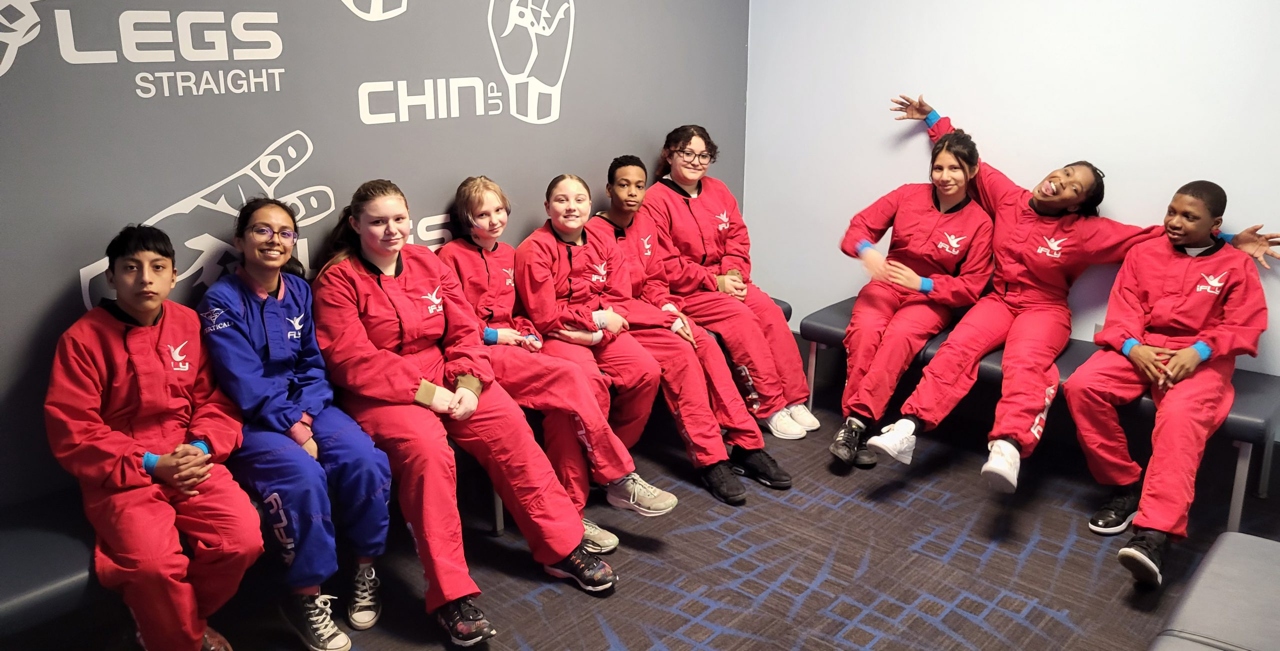 Students pose for a photo seated on benches and suited up for the wind tunnel