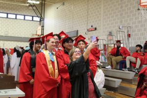 A teacher poses for a selfie with a group of graduating seniors in their caps and gowns