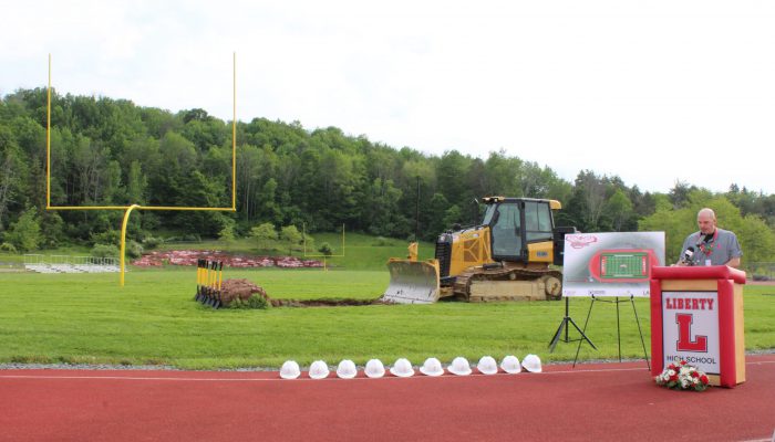 groundbreaking10 A man speaks at a podium with Liberty and a block L with a board featuring the design of the new field and track to his right as well as a bulldozer on the field and a line of white hard hats on the track.