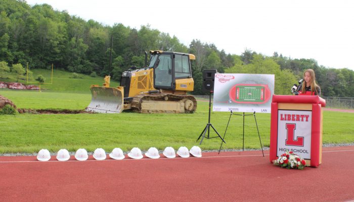 groundbreaking6 A student speaks at a podium with Liberty and a block L with a board featuring the design of the new field and track to her right as well as a bulldozer on the field and a line of white hard hats on the track.
