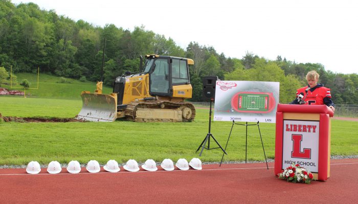 groundbreaking3 A student speaks at a podium with Liberty high school on it with a rendering of the athletic field and a bulldozer behind him and a line of white hard hats on the ground to his right.