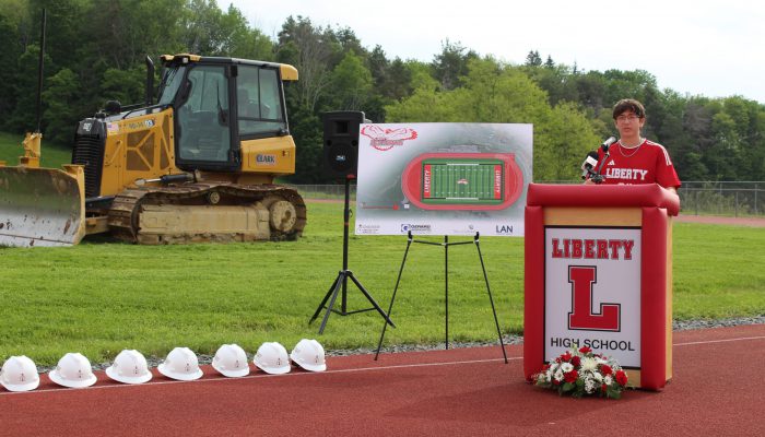groundbreaking4 A student speaks at a podium with Liberty and a block L with a board featuring the design of the new field and track to his right as well as a bulldozer on the field and a line of white hard hats on the track.
