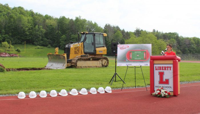 Groundbreaking 5 A student speaks at a podium with Liberty and a block L with a board featuring the design of the new field and track to his right as well as a bulldozer on the field and a line of white hard hats on the track.