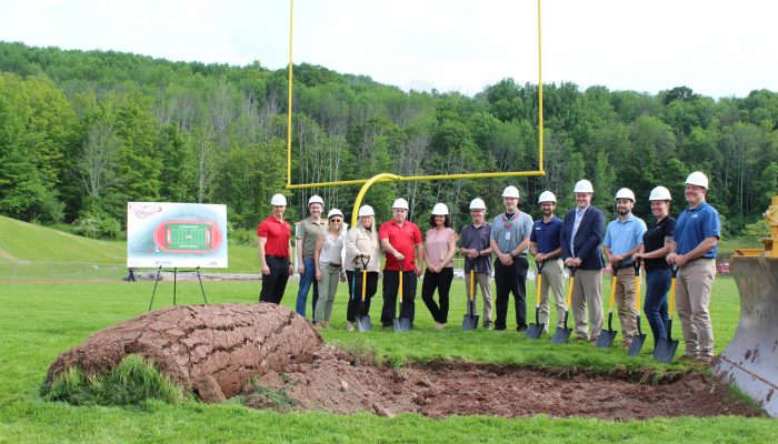 groundbreaking8 Administrators and officials pose with hard hats next to a pile of dirt and grass with a board featuring the new field and track next to them.