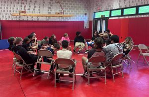 Students and adults sit in a circle in chairs on a red mat in a gym