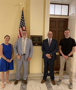 Four people stand in front of an office with an American flag on the other side of the wall from the door