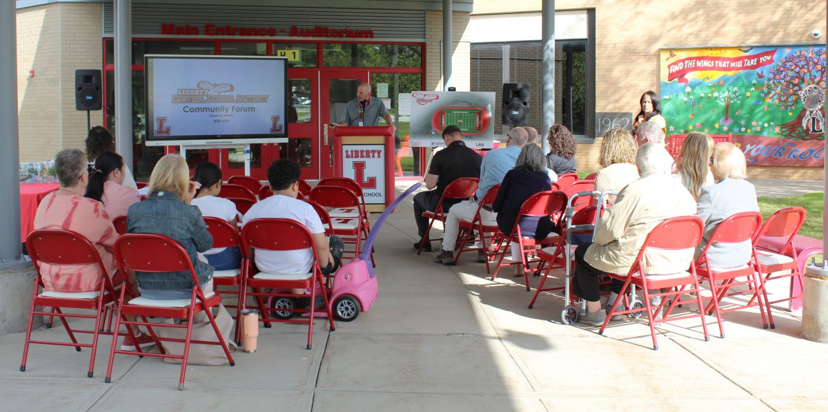 A man speaks at a podium with a large screen to this right as an audience watches