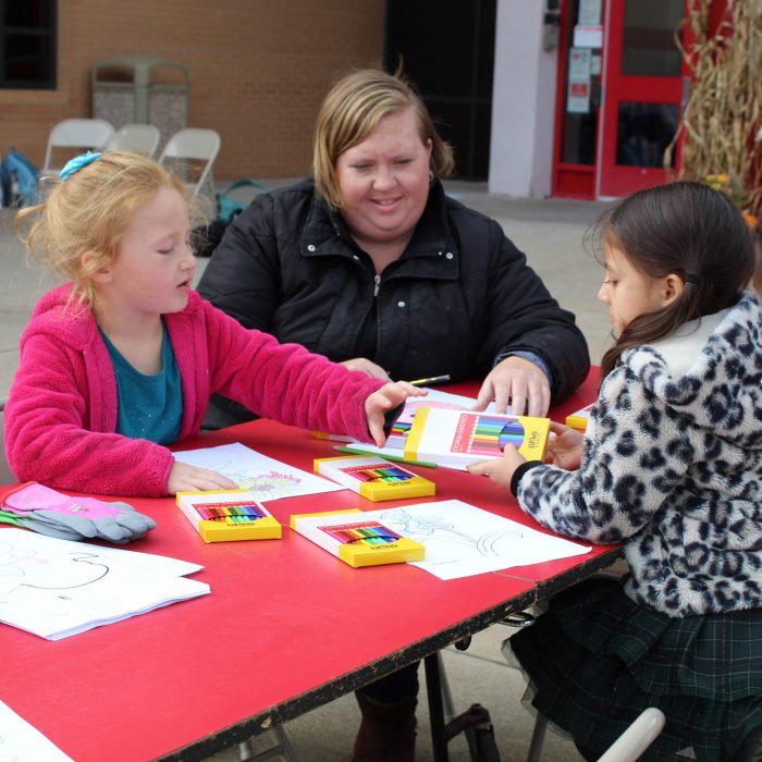 A student hands colored pencils to another student as an adult watches