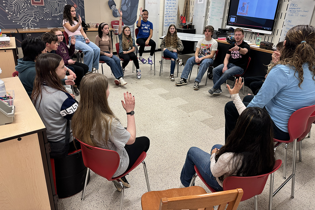 Students and others sit in chairs in a circle.