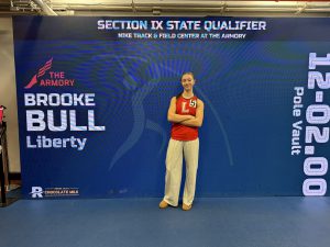 A Liberty student poses in front of a blue screen with Section IX State Qualifiers. and Brook Bull Liberty on it.