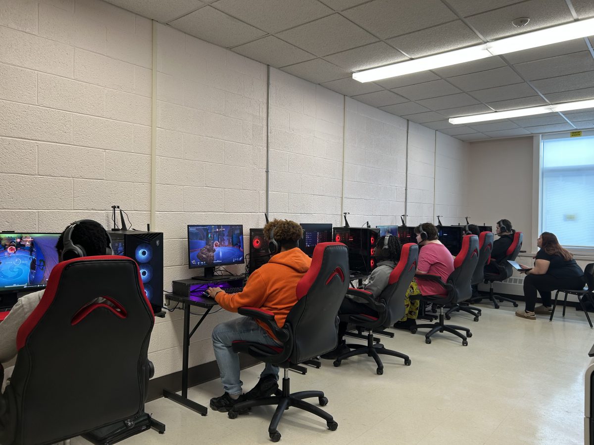 Six players siting at PC gaming stations compete as a team in Marvels Rivals as a coach watches.