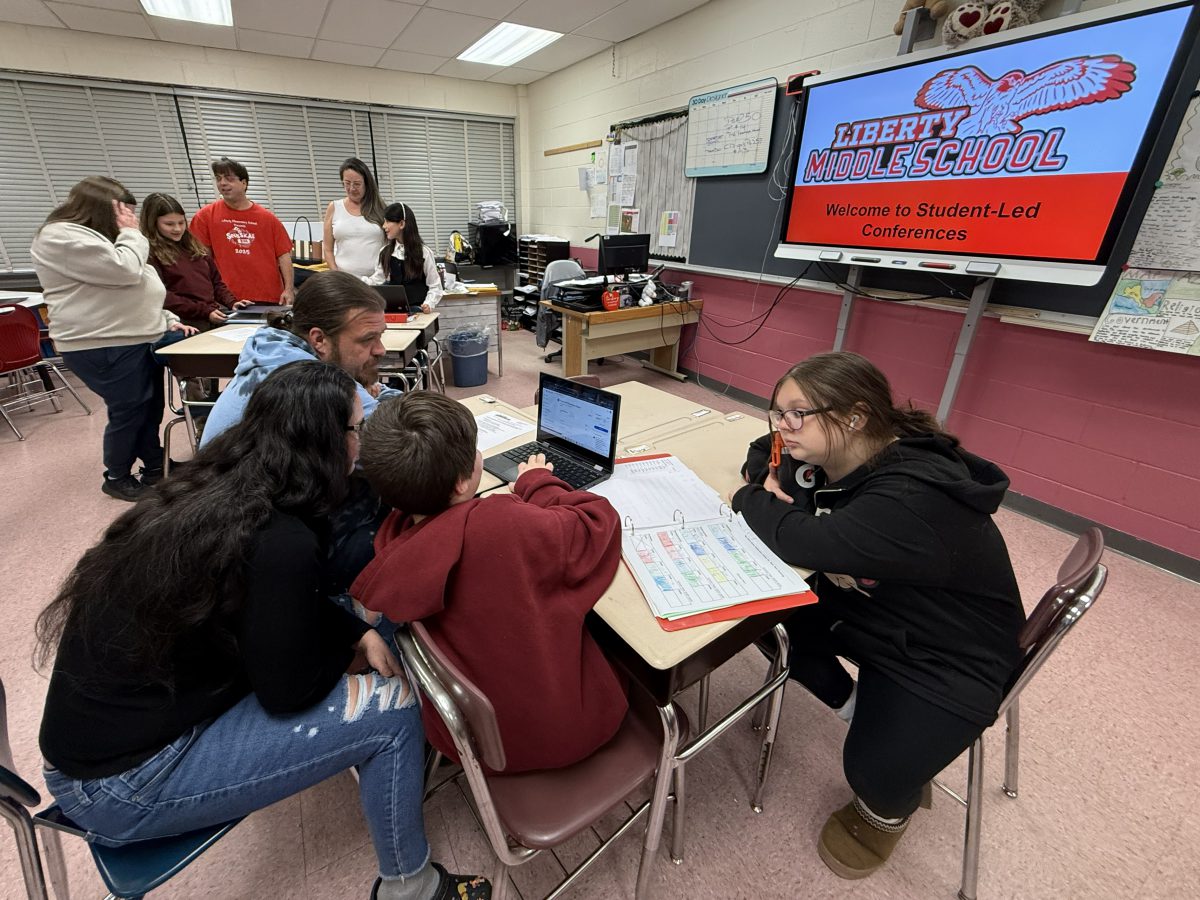 Students and adults sit at tables with Student Led Conferences on a screen behind them.